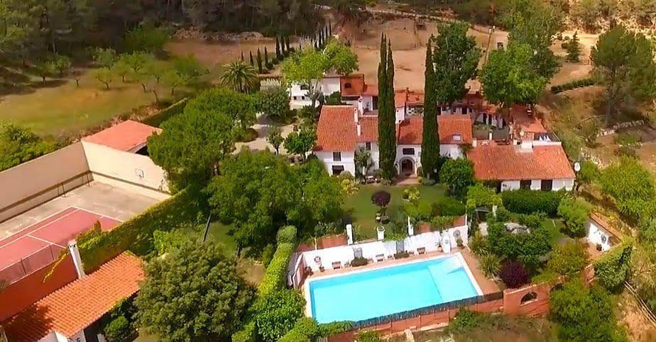 An aerial view showcases a rustic property surrounded by lush greenery and tall trees, featuring terracotta-roofed buildings. A rectangular swimming pool is visible in the center, complemented by sun loungers. Nearby, a tennis court is framed by gardens and natural landscape.