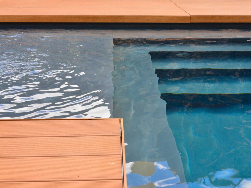 The image features a swimming pool with clear, inviting water. Steps leading into the pool are visible, alongside a wooden deck that borders the edge. The reflections on the water's surface enhance the serene ambiance of the area.