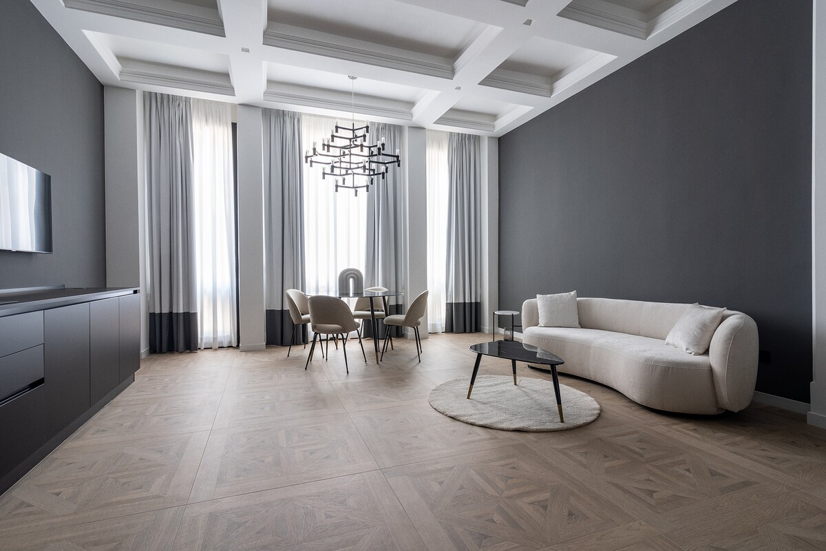 An open living area is featured, showcasing a modern curved sofa and a round coffee table on a textured rug. A dining table with four chairs is situated beside large windows dressed in long curtains. A contemporary chandelier hangs from a detailed ceiling.