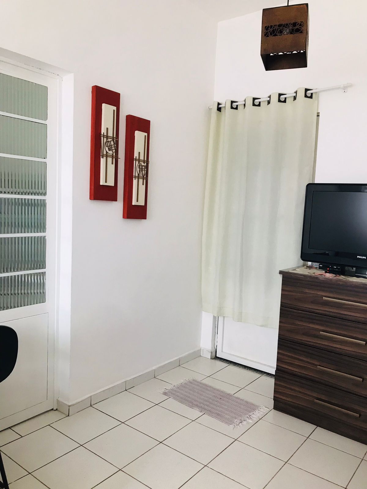 A bright corner of the kit features a sliding glass door covered by a light, sheer curtain. Two framed artworks in red and cream tones hang on the wall above a sleek wooden dresser. A small television is placed on top of the dresser.