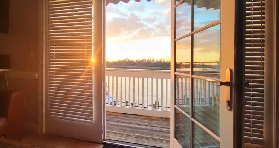 A view from inside the apartment features open glass doors leading to a balcony. A golden sunset illuminates the scene, highlighting the serene waters of the river below. White railings can be seen on the balcony, enhancing the tranquil setting.
