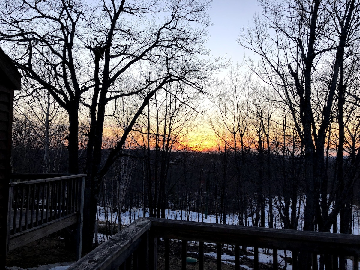 A serene sunset is captured through silhouetted trees, revealing a gradient of orange and purple tones in the sky. A wooden deck railing is visible in the foreground, providing a sense of depth to the tranquil natural setting.