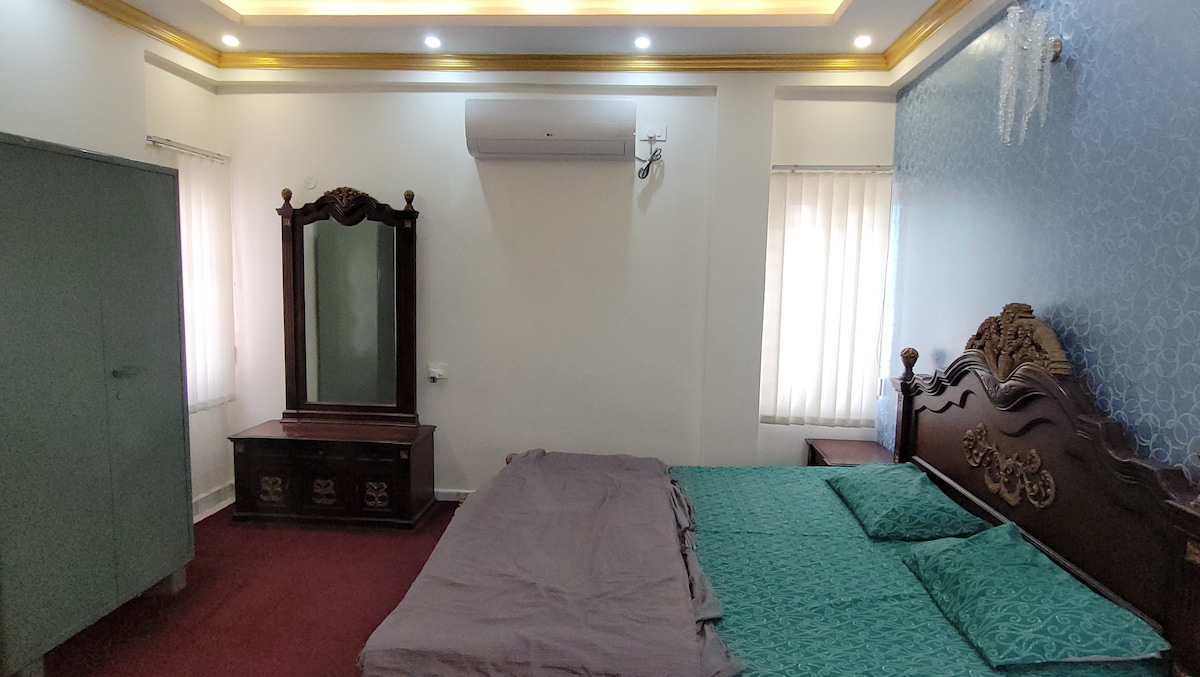 A spacious bedroom is equipped with a large bed featuring decorative headboards and green bedding. A wooden dresser with a mirror is positioned against the wall. The room is illuminated by soft ceiling lights, and natural light filters through sheer curtains on the windows.