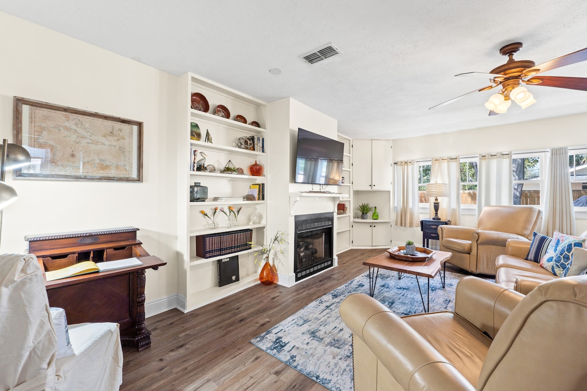 A comfortable living area is furnished with two light-colored armchairs and a small wooden table. Built-in shelves display various decorative items. A television is mounted above a fireplace, while large windows allow natural light to fill the space.