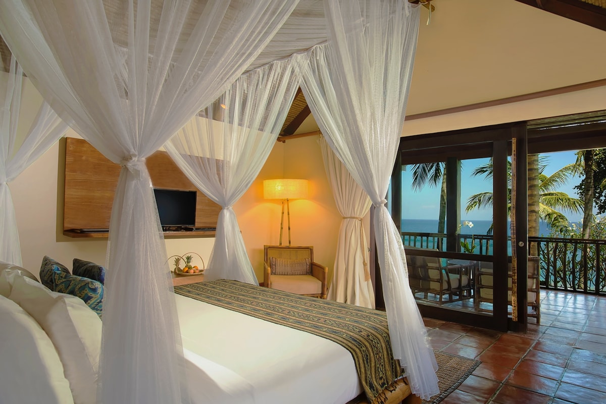 An air-conditioned suite features a spacious bed with draped white curtains. A soft lighting fixture beside the bed enhances the tranquil ambiance. Glass doors open to a terrace, providing views of the lush palm trees and the ocean beyond.