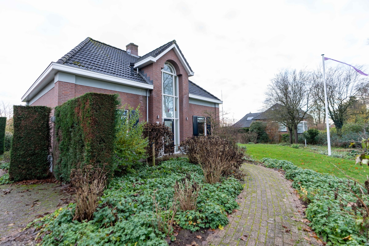 The exterior of a charming brick house is showcased, framed by neatly trimmed hedges and seasonal shrubs. A winding path leads to the entrance, while the surrounding garden is home to various plants and trees, contributing to a serene and inviting environment.