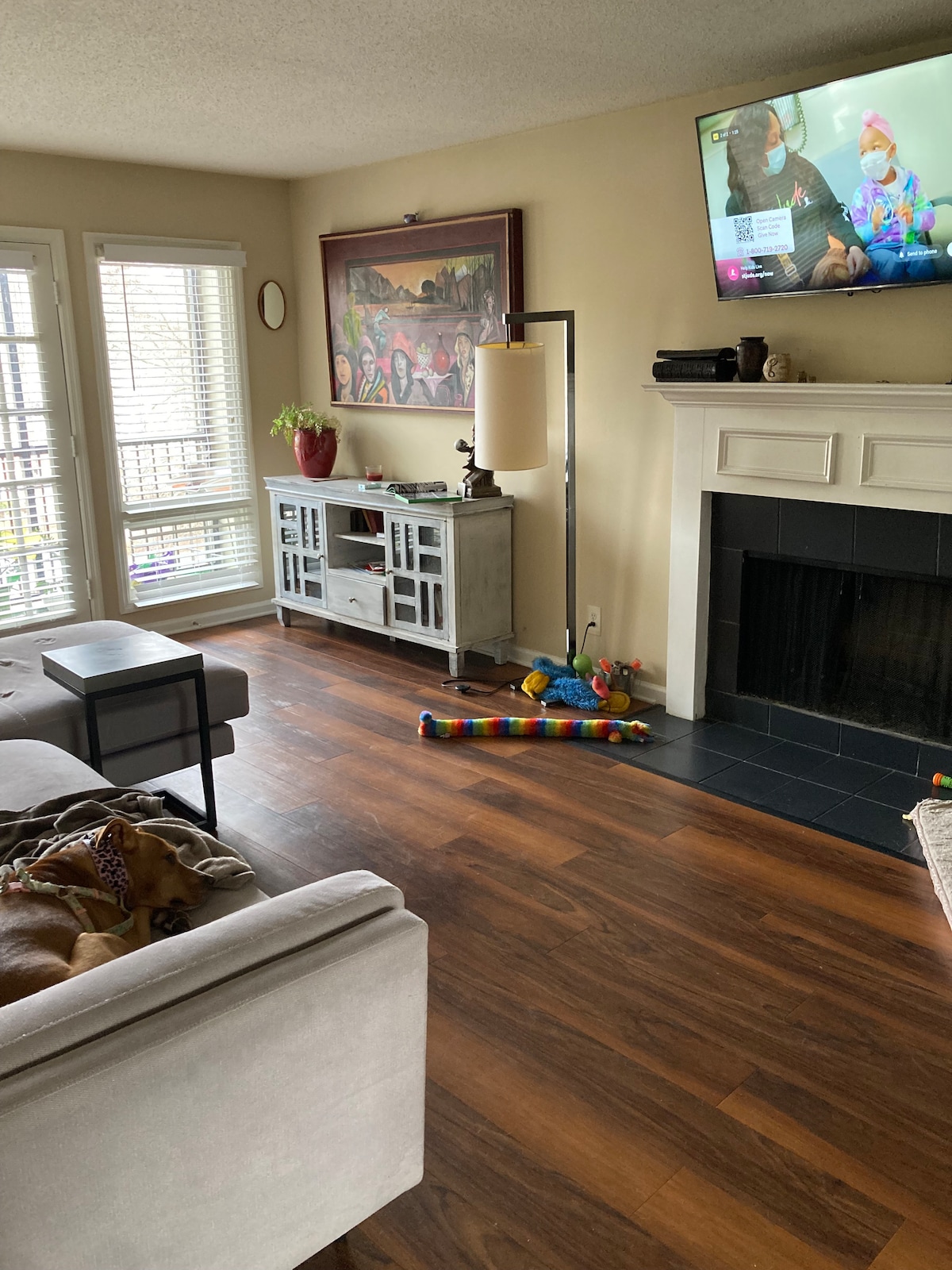 A spacious living area features a comfortable light-colored couch and a coffee table set atop warm wooden flooring. A television is mounted above a fireplace, while a large artwork adorns the wall. Natural light filters through the sliding glass doors, enhancing the open atmosphere.