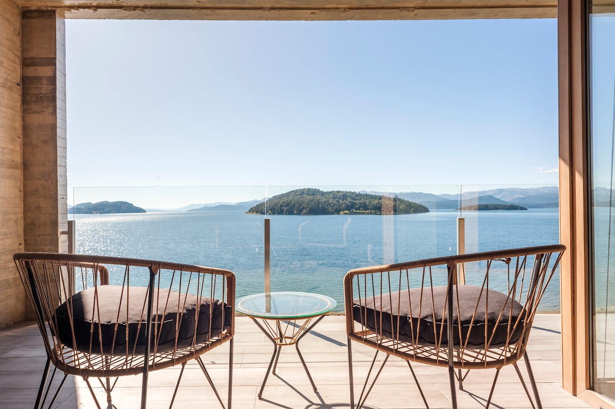 A private terrace is equipped with two woven chairs and a glass table, offering unobstructed views of the lake and surrounding islands. The bright blue sky complements the serene water, creating a calming outdoor space for relaxation.