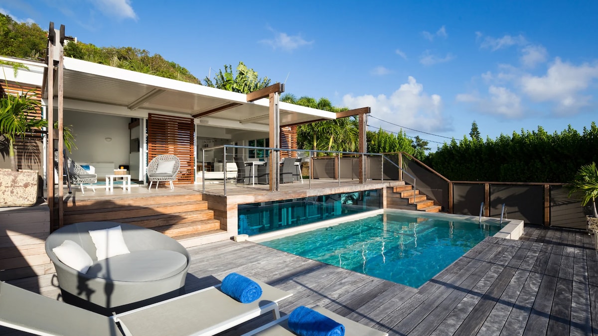 An outdoor area is showcased, featuring a tranquil swimming pool surrounded by wooden decking. Lounge chairs equipped with blue towels are positioned for relaxation. A covered terrace is visible in the background, offering space for seating and enjoying the surrounding greenery.