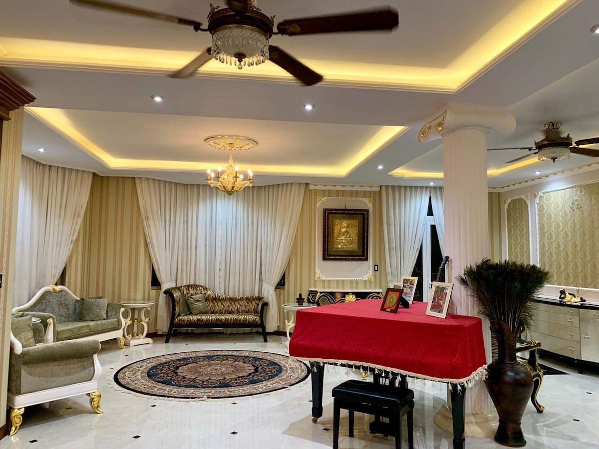 An elegant living area is presented with a red table at its center, surrounded by plush seating. Soft lighting illuminates the room, highlighting decorative wall panels and a round area rug. Large windows are draped with light curtains, enhancing the spacious ambiance.