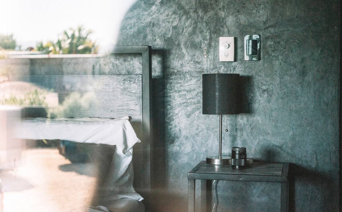 A spacious bedroom features a textured gray wall and a cozy bed with crisp white linens. A bedside table holds a modern lamp and a small container. Natural light streams through a nearby window, illuminating the serene space.