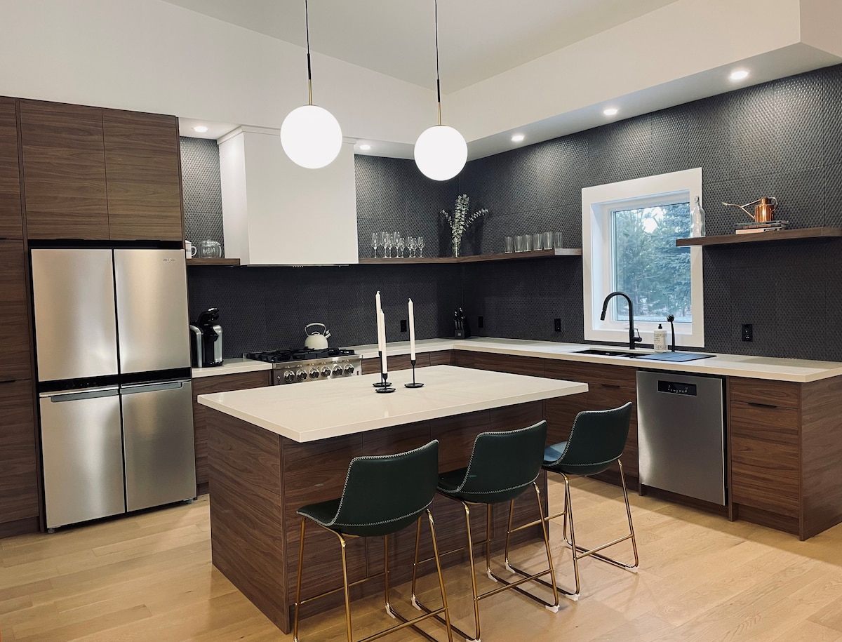 A modern kitchen features sleek wooden cabinetry and stainless steel appliances. An island with a light countertop offers seating with three green chairs. Soft lighting from two pendant lights enhances the space, and a window provides natural light over the sink area.