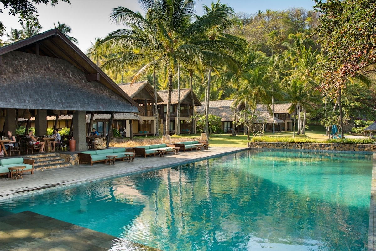 A serene swimming pool invites relaxation, bordered by gentle palm trees and lush gardens. Traditional architecture is visible in the background, with sun loungers positioned near the water's edge, offering a tranquil setting for guests.