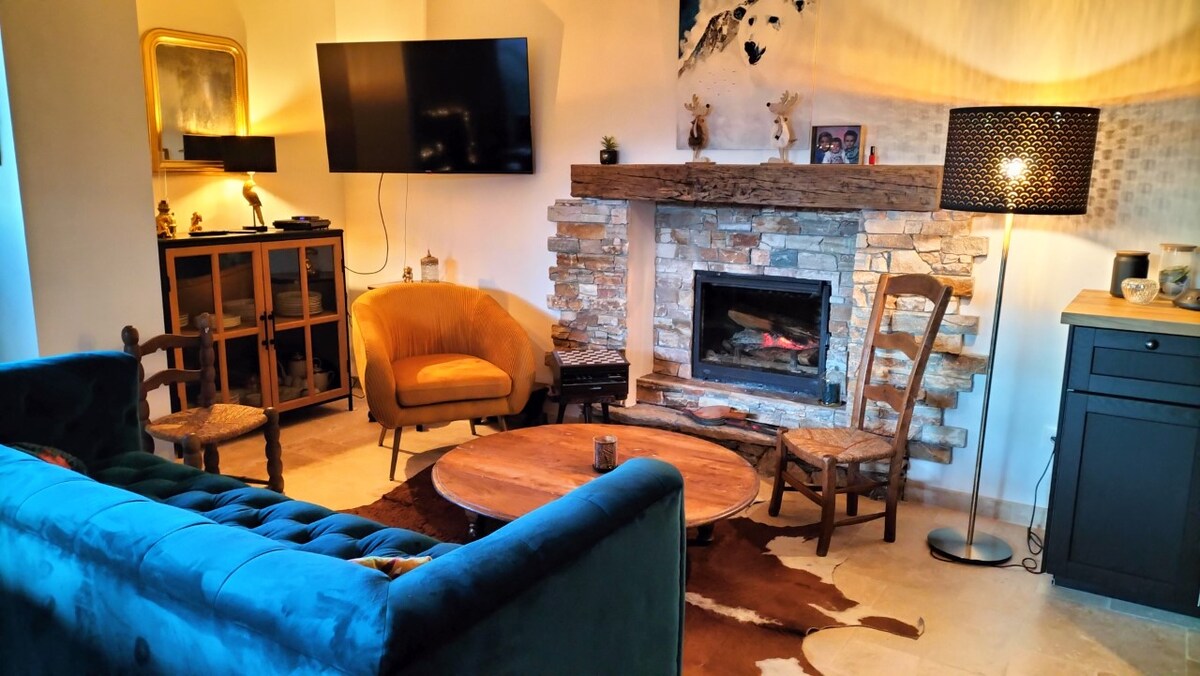 A cozy living area features a blue tufted sofa and an orange armchair, accompanied by wooden chairs. A rustic stone fireplace serves as a focal point, with a wall-mounted television above. Soft lighting is provided by a floor lamp, enhancing the warm ambiance.