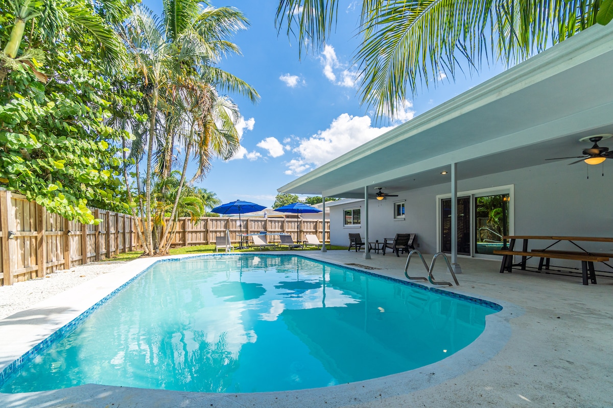 A bright outdoor area features a refreshing pool surrounded by light concrete flooring. Lush palm trees provide shade, while a picnic table sits nearby. An umbrella offers additional outdoor seating options, creating a serene space for relaxation amid a private fenced yard.