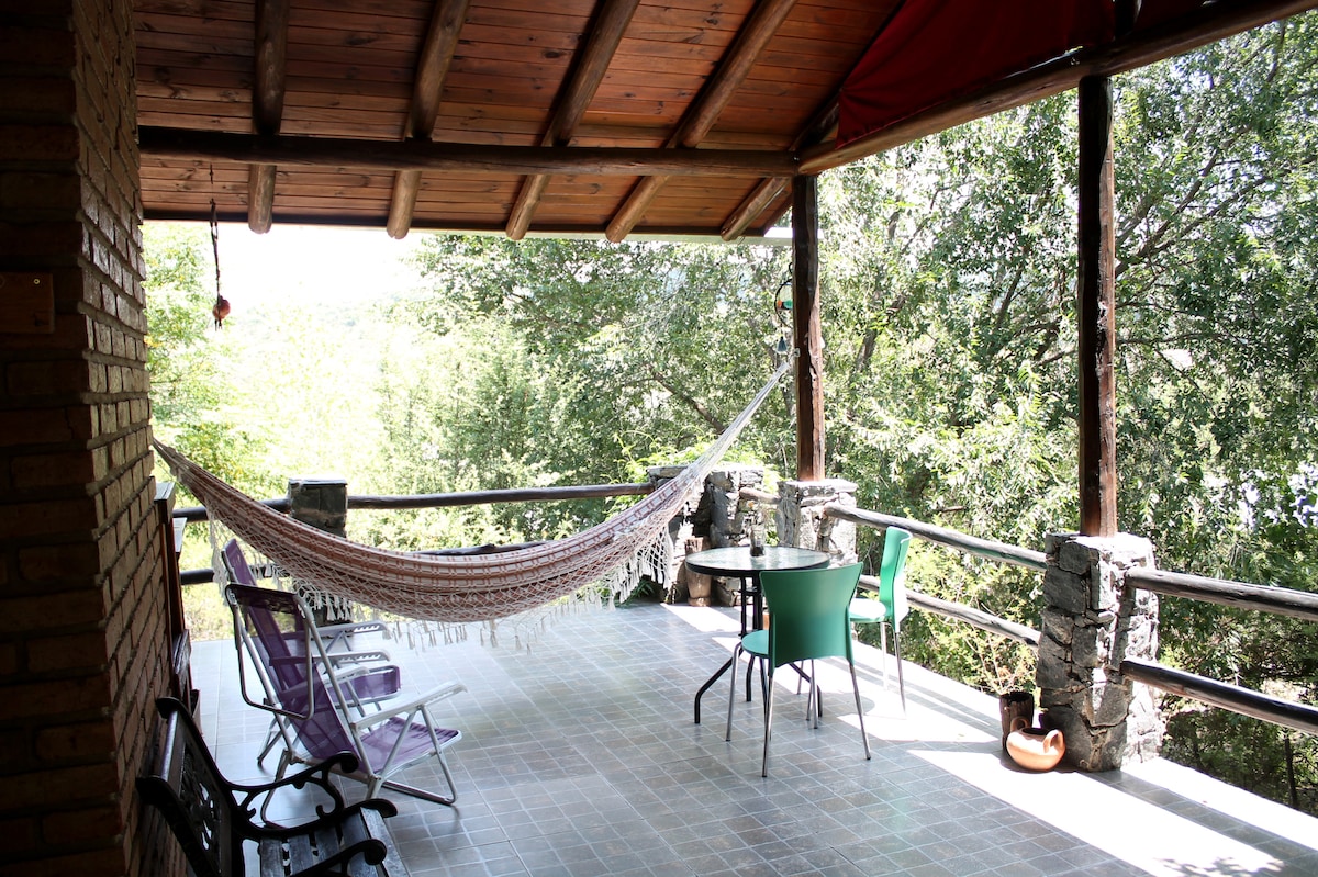 An outdoor gallery features a comfortable hammock and a table set for four, surrounded by a railing offering views of lush greenery. Two chairs and a bench are positioned nearby, inviting relaxation amidst a serene natural environment.