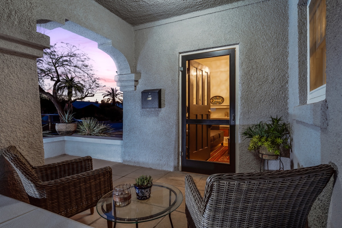 An outdoor patio area features two wicker chairs placed around a glass-topped table, adorned with a small potted plant. The entrance is visible through a glass door, with warm lighting creating an inviting glow as dusk settles in the background.
