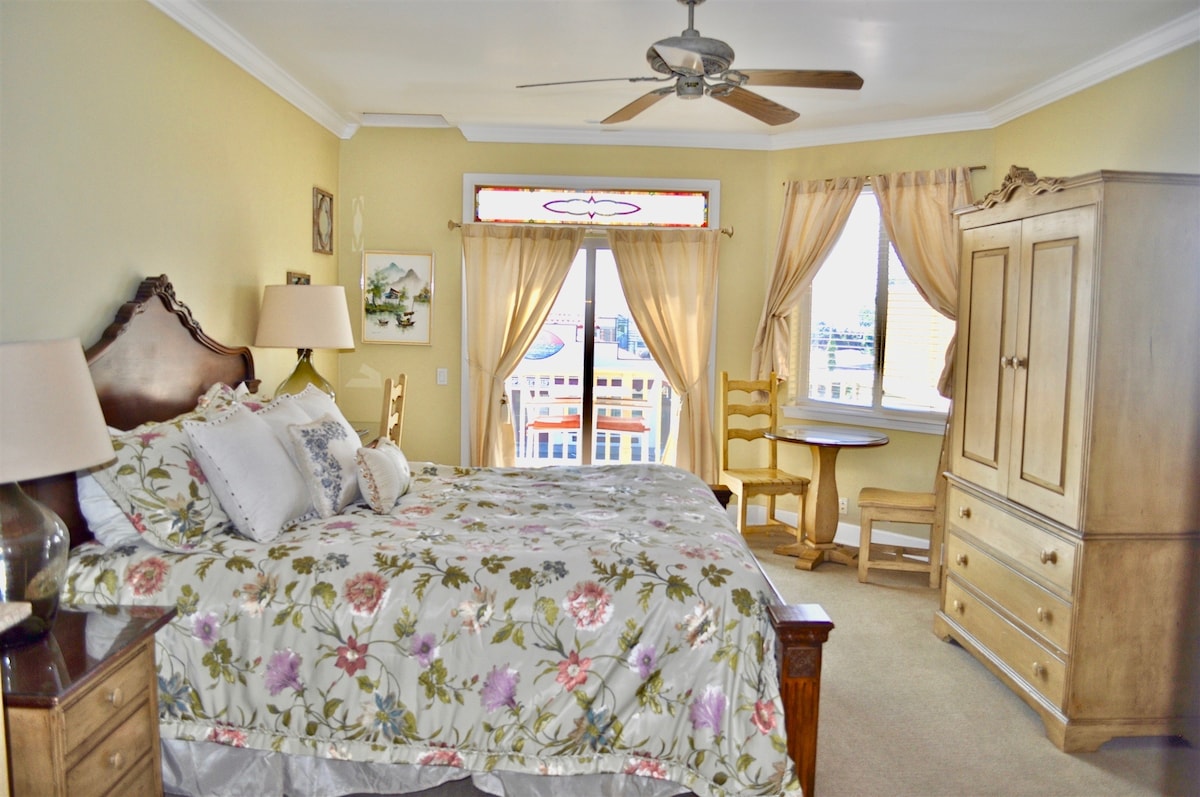 A spacious bedroom features a large bed adorned with a floral-patterned quilt. Natural light enters through a window with sheer curtains, illuminating the room. A wooden dresser and a small table with a chair are positioned nearby, enhancing the room's cozy and inviting ambiance.
