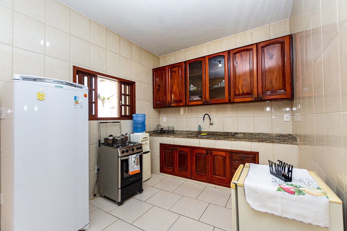 The kitchen appears spacious and features wooden cabinetry and a granite countertop. A stove and a refrigerator are present, along with a water dispenser. Natural light enters through a window above the sink, illuminating the tiled walls and floor.