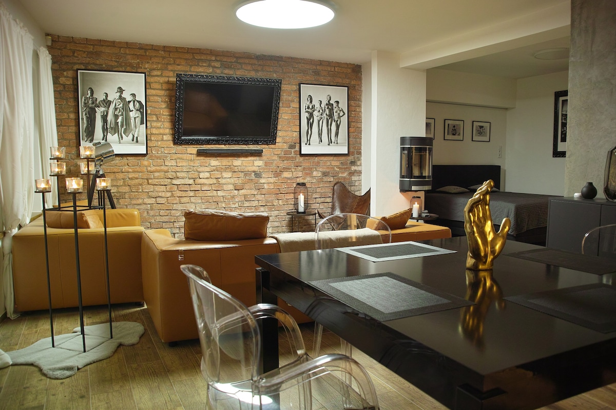 A stylish living area features a mix of warm-toned furniture, including two leather sofas and clear plastic chairs. A dark wooden dining table sits prominently, accompanied by a decorative gold sculpture. A large wall-mounted TV and framed artwork enhance the modern design.