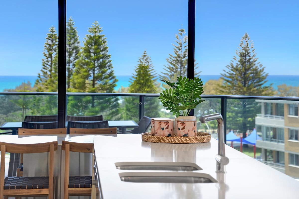 A modern kitchen area features a sleek white countertop with a double sink. Natural light floods the space through large windows, offering stunning ocean views and surrounded by greenery. A decorative plant and two patterned containers are displayed on the counter, enhancing the inviting atmosphere.