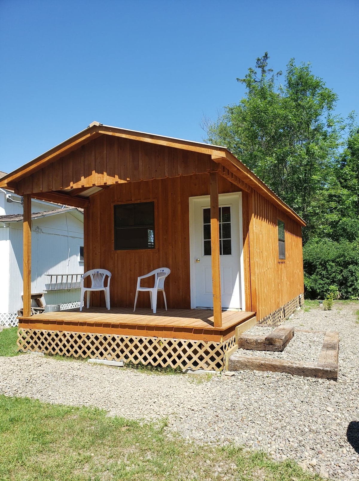 A simple wooden cabin with a welcoming front porch, featuring two white chairs. The cabin's exterior is enhanced by natural wood tones and a sloped roof. Lush greenery is visible in the background, contributing to the overall serene environment.