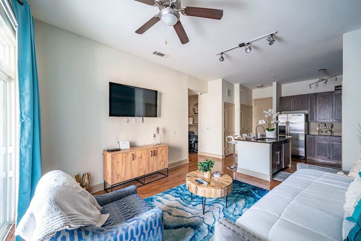 The living room features a smart TV mounted on the wall, a stylish coffee table surrounded by a comfortable light blue chair, and a cozy daybed. Large windows allow natural light to fill the space, complemented by a vibrant area rug and modern decor.