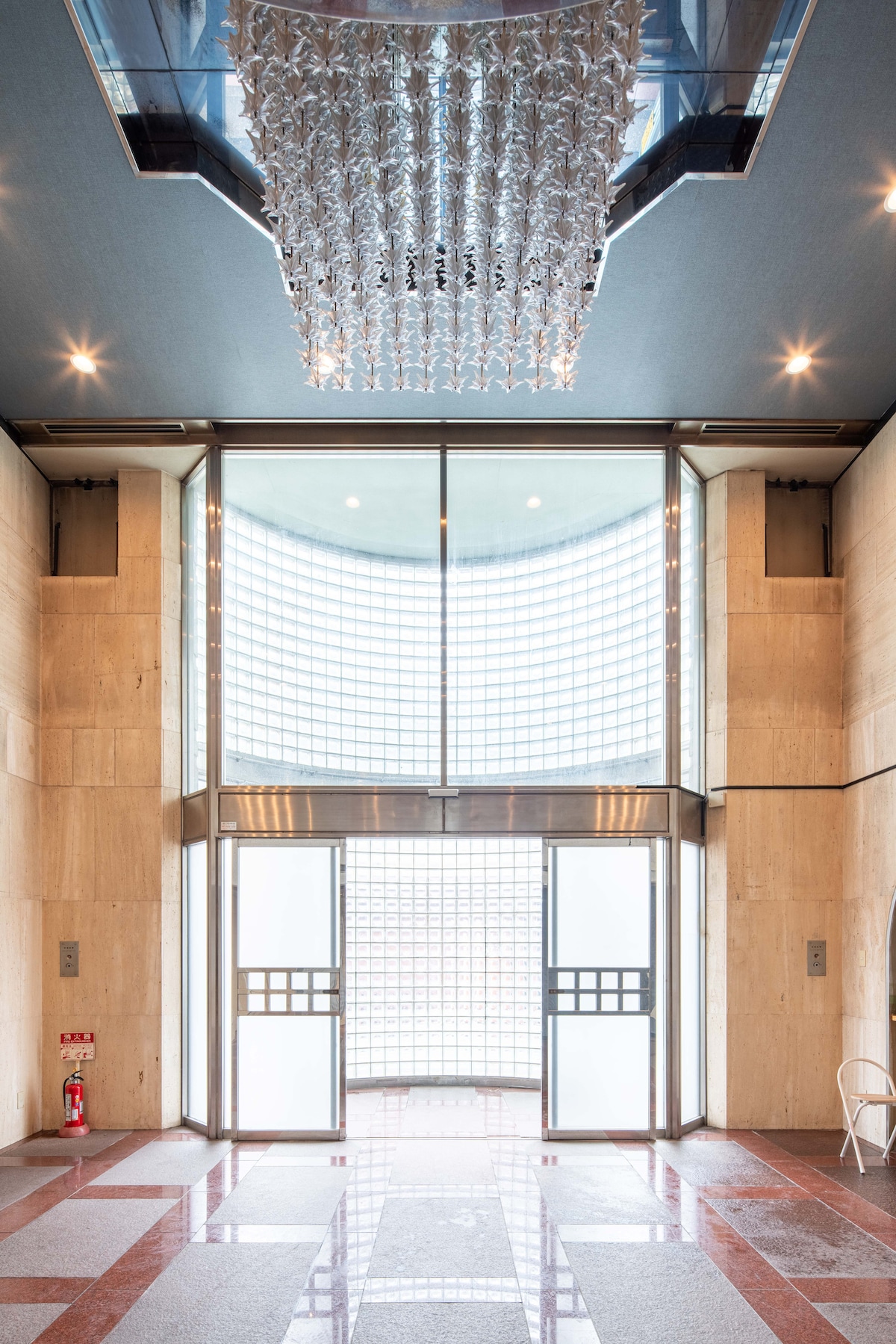 A spacious entrance is illuminated by natural light filtering through a large glass wall. The sleek, modern design features decorative lighting overhead and polished floor tiles. Dual glass doors provide an inviting entry point, complemented by minimalist elevator panels on either side.