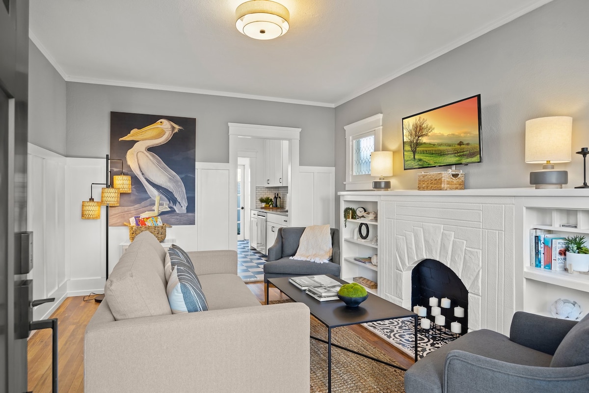 The living area is arranged with a comfortable sofa facing a sleek coffee table. A large artwork of a bird hangs above the mantel, complemented by soft lighting from table lamps. The fireplace features a white surround, and a glimpse of the kitchen is visible through the door.