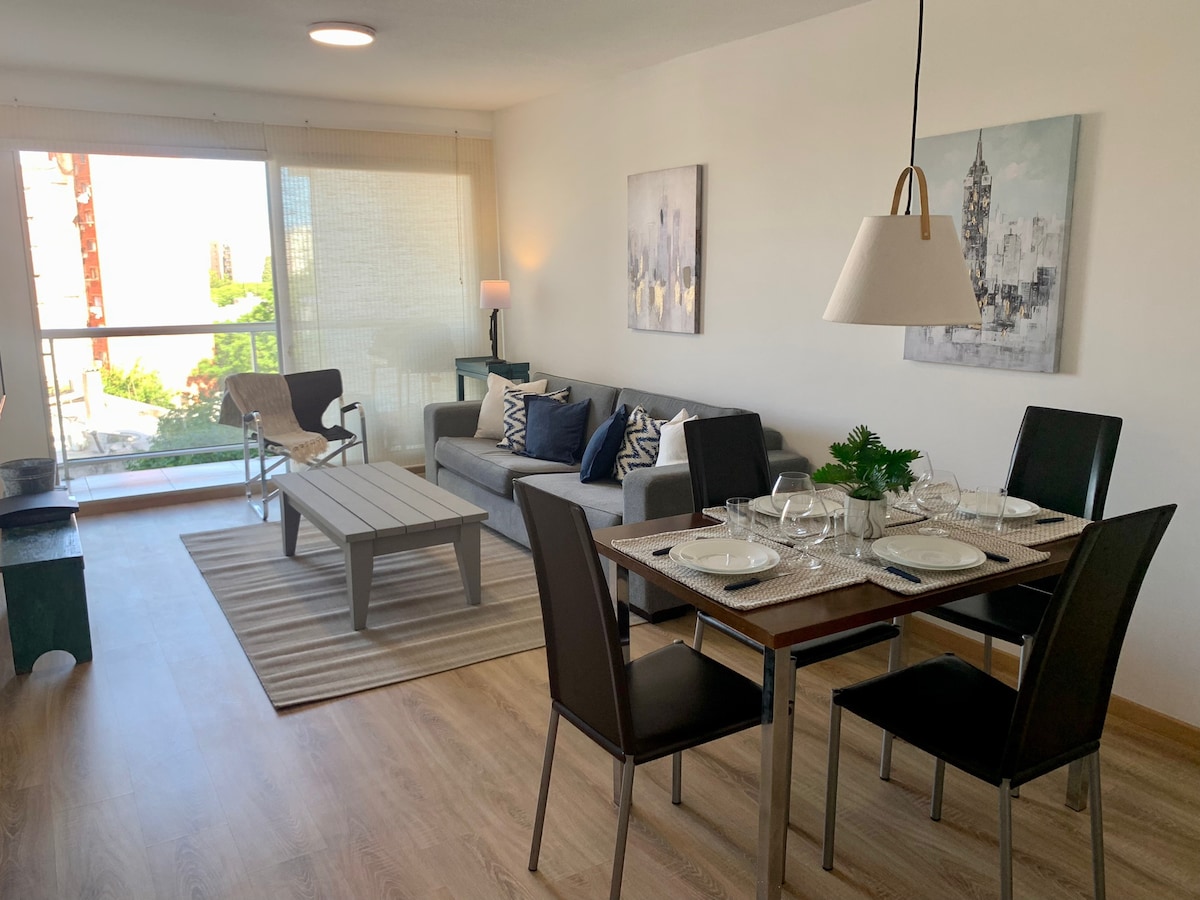 A modern living space features a comfortable sofa adorned with patterned cushions, accompanied by a dining area with a polished table set for four. Large windows provide natural light, with outdoor views visible through sheer curtains.