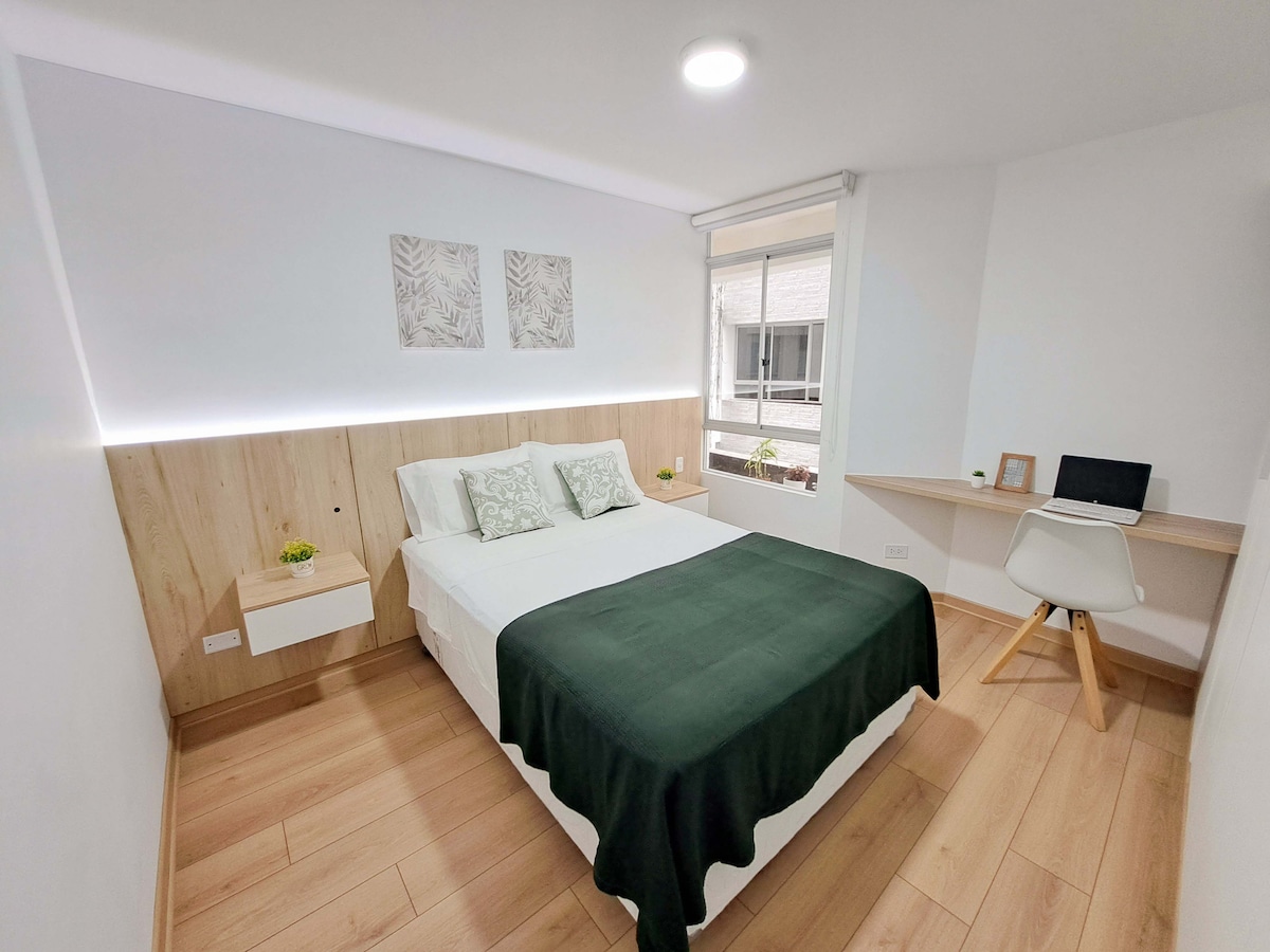 A well-lit bedroom features a spacious bed with a dark green blanket. Two decorative wall pieces hang above the bed, complementing the light wooden accents. A comfortable workspace is situated near the window, providing a serene view of the outside.