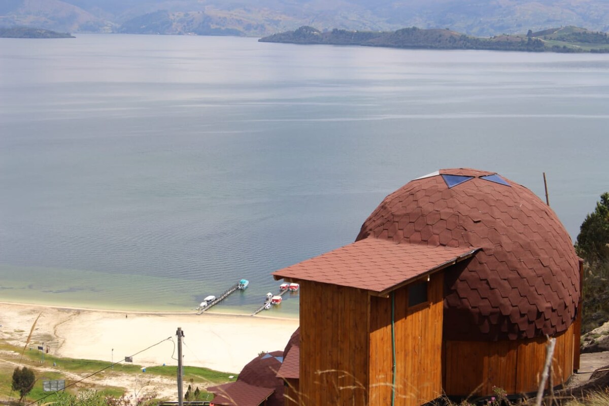 A unique glamping structure with a rounded roof is situated on a hillside, overlooking a tranquil lake. The sandy beach is visible below, along with several boats docked at a nearby pier, framed by distant mountains under a clear sky.