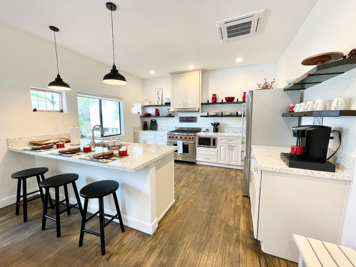 The kitchen features modern appliances and elegant cabinetry, complemented by a spacious granite countertop. A coffee station is visible, alongside thoughtfully arranged tableware. Two black stools are positioned at the kitchen counter, providing an inviting area for casual dining or conversation.