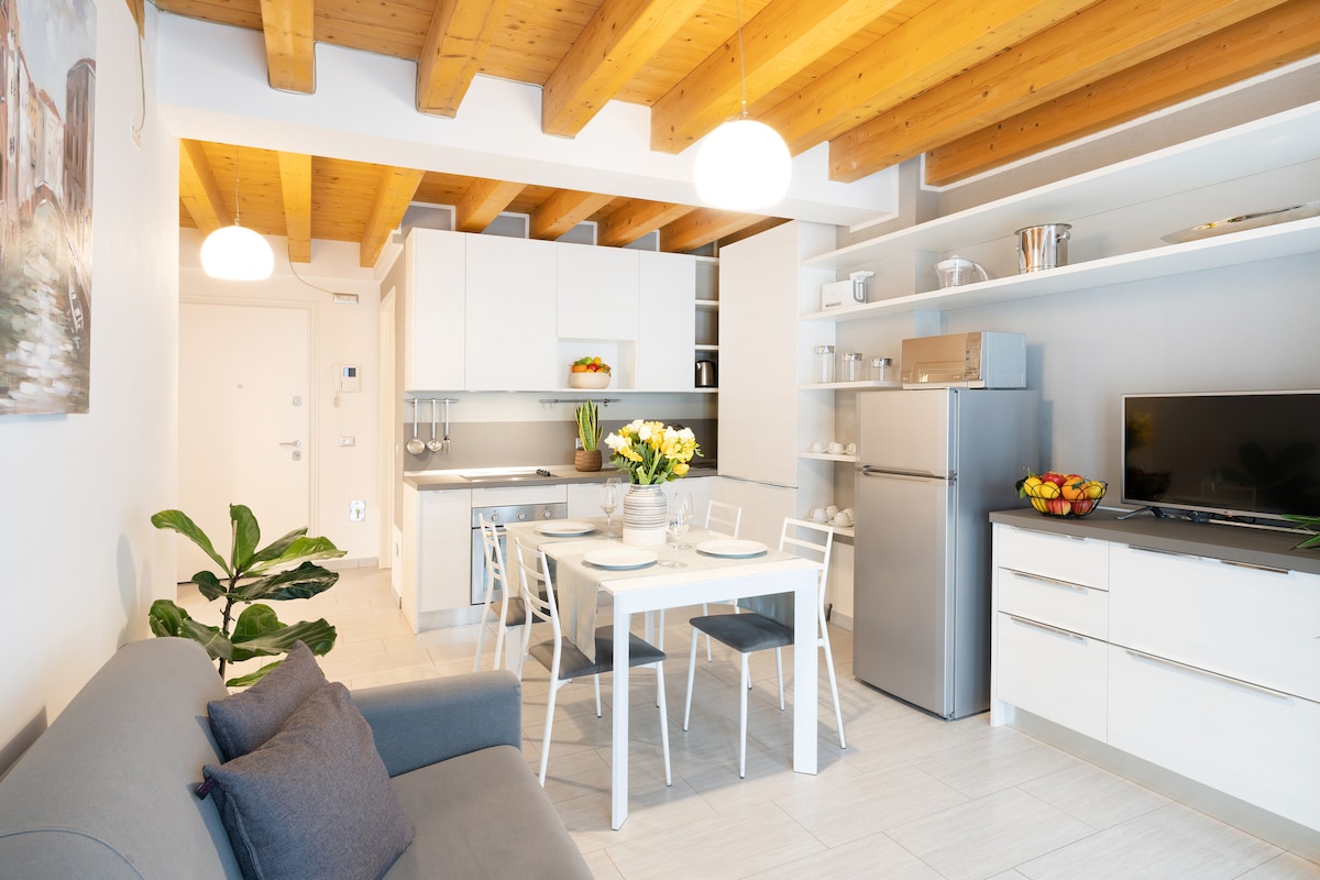 A modern kitchen and living area are presented, featuring an open layout with white cabinetry and a stainless-steel refrigerator. A dining table with four chairs is positioned near a window, complemented by a comfortable sofa adorned with cushions. Light fixtures hang from the wooden ceiling beams.