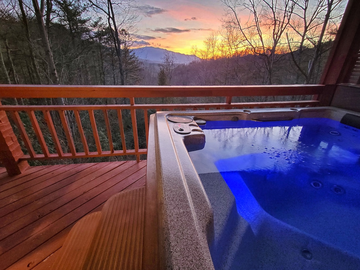 A relaxing hot tub is positioned on a wooden deck, with the calming water reflecting soft blue lights. Beyond, the sun sets over a scenic mountain range, framed by bare trees, creating a tranquil atmosphere for unwinding in nature.