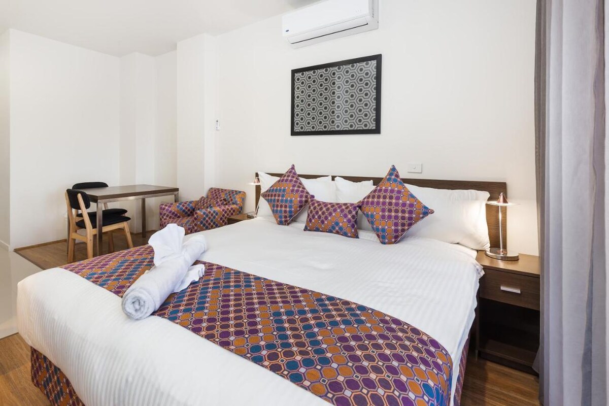 A spacious bedroom features a large bed adorned with decorative pillows and a colorful throw blanket. Side tables with lamps are placed on either side. A desk and chair are visible in the background, with a wall art piece adding character to the space.