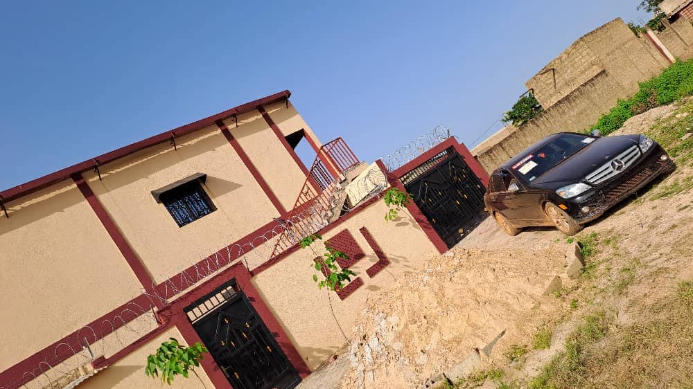 The exterior of the building features a two-story structure with a red and beige color scheme. A black car is parked adjacent to a sandy area, with plants growing nearby. A staircase leads to the upper level, and security features are visible.