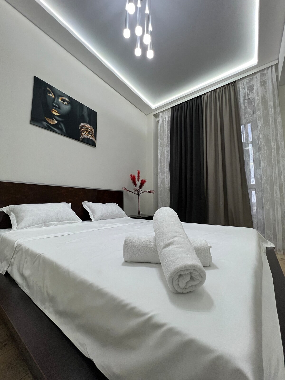 A spacious sleeping area is furnished with a large bed topped with fresh white linens. Two rolled towels are placed neatly on the bed. Soft, sheer curtains frame a window, while a subtle piece of artwork adorns the wall above the bed, adding a touch of elegance.