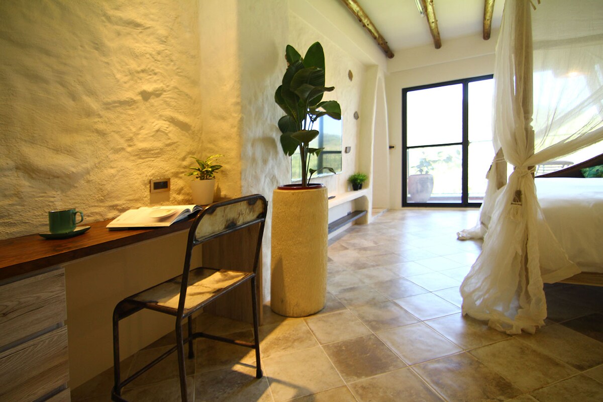 A serene room features a rustic desk with a chair and a small potted plant. Natural light fills the space through glass doors that lead outside. A spacious bed is visible, complemented by soft, sheer drapery and an earthy tiled floor.