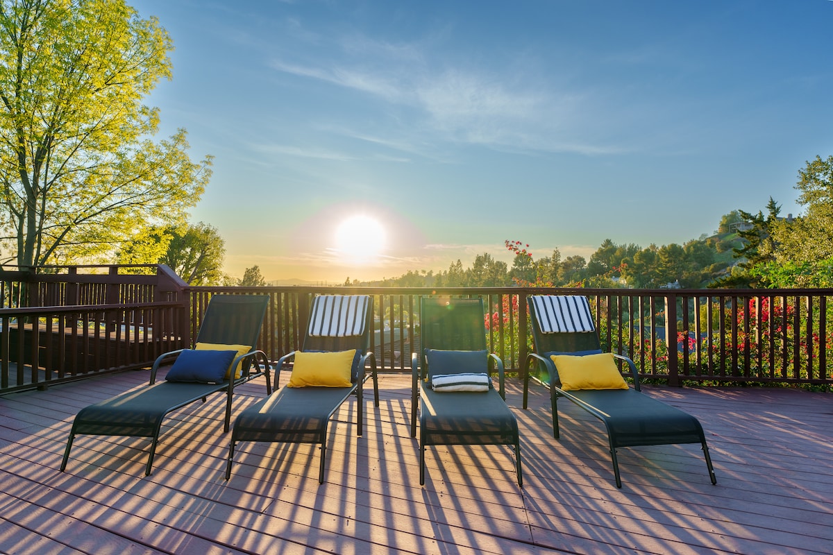 A spacious outdoor deck is equipped with four sun loungers, each featuring a yellow accent pillow. The scene captures a sunset over a lush landscape, with trees and colorful plants visible in the background.