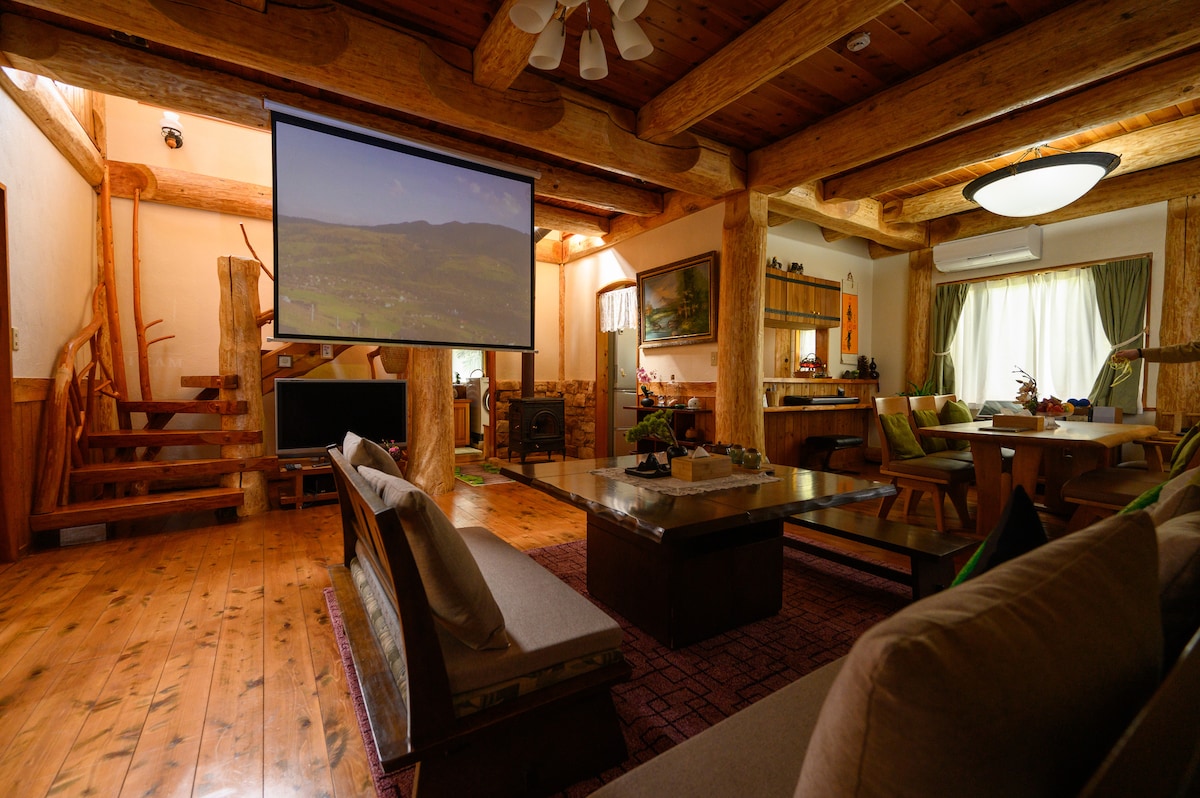 An inviting living area features wooden beams and warm lighting, complemented by a large screen mounted on the wall. A spacious coffee table sits at the center, surrounded by comfortable seating. The adjacent kitchen area is visible, showcasing a blend of natural elements and modern comforts.