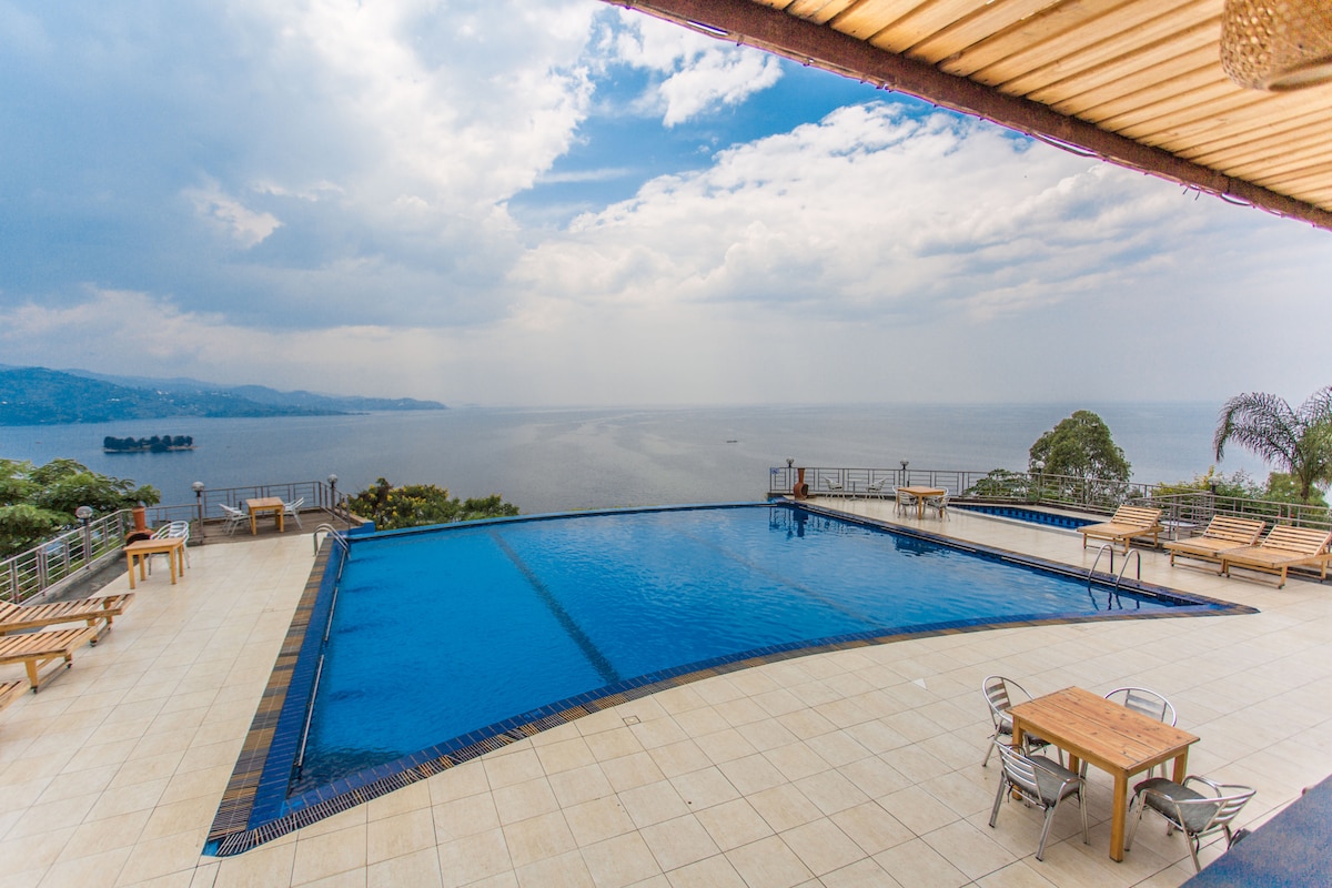 An expansive infinity pool reflects the sky, overlooking Lake Kivu. Several lounge chairs are arranged around the pool on a tiled deck, complemented by wooden tables. Lush greenery frames the area, enhancing the serenity of the scenic view of the water and distant mountains.