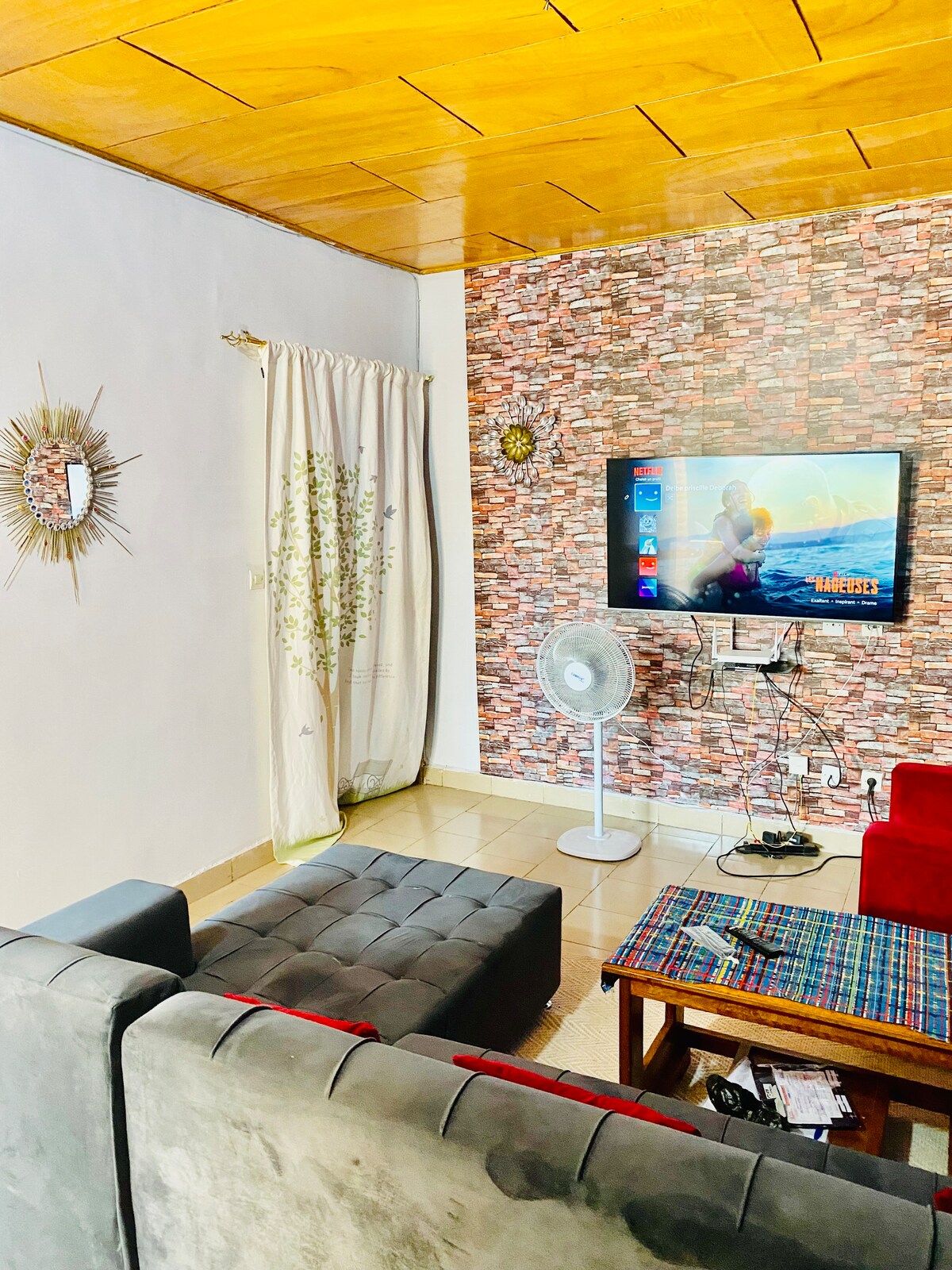 A cozy living area features a grey sofa and a red armchair, complemented by a textured coffee table. A wall-mounted television is positioned above a fan, and a patterned throw is placed on the seating. Natural light enters through a curtain adorned with botanical prints.