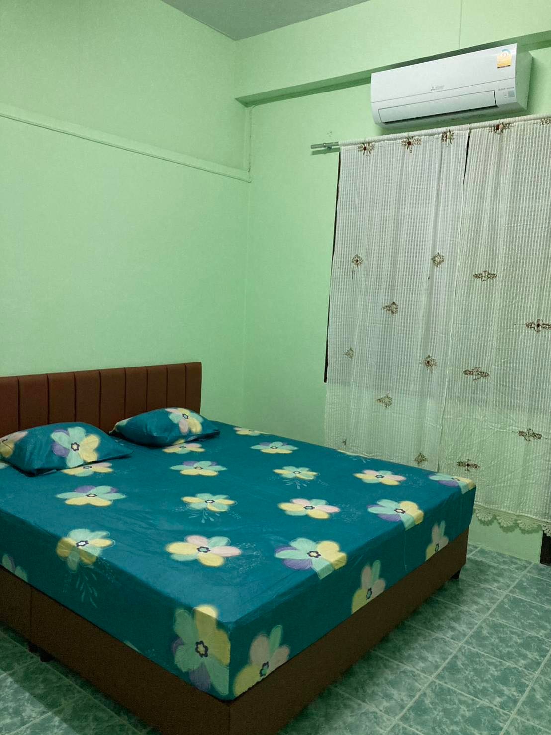 A cozy sleeping area features a spacious bed dressed in a vibrant blue floral sheet. Two matching pillows are positioned at the head of the bed. Natural light enters through a partially sheer curtain, and a wall-mounted air conditioning unit is visible above.