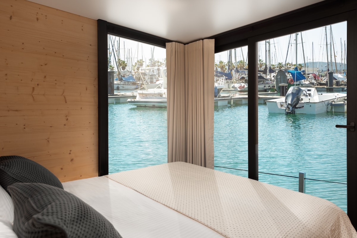 A spacious bedroom features large windows with stunning views of the marina and boats anchored in the serene water. Soft, neutral bedding is complemented by two plush pillows, creating a calming atmosphere.