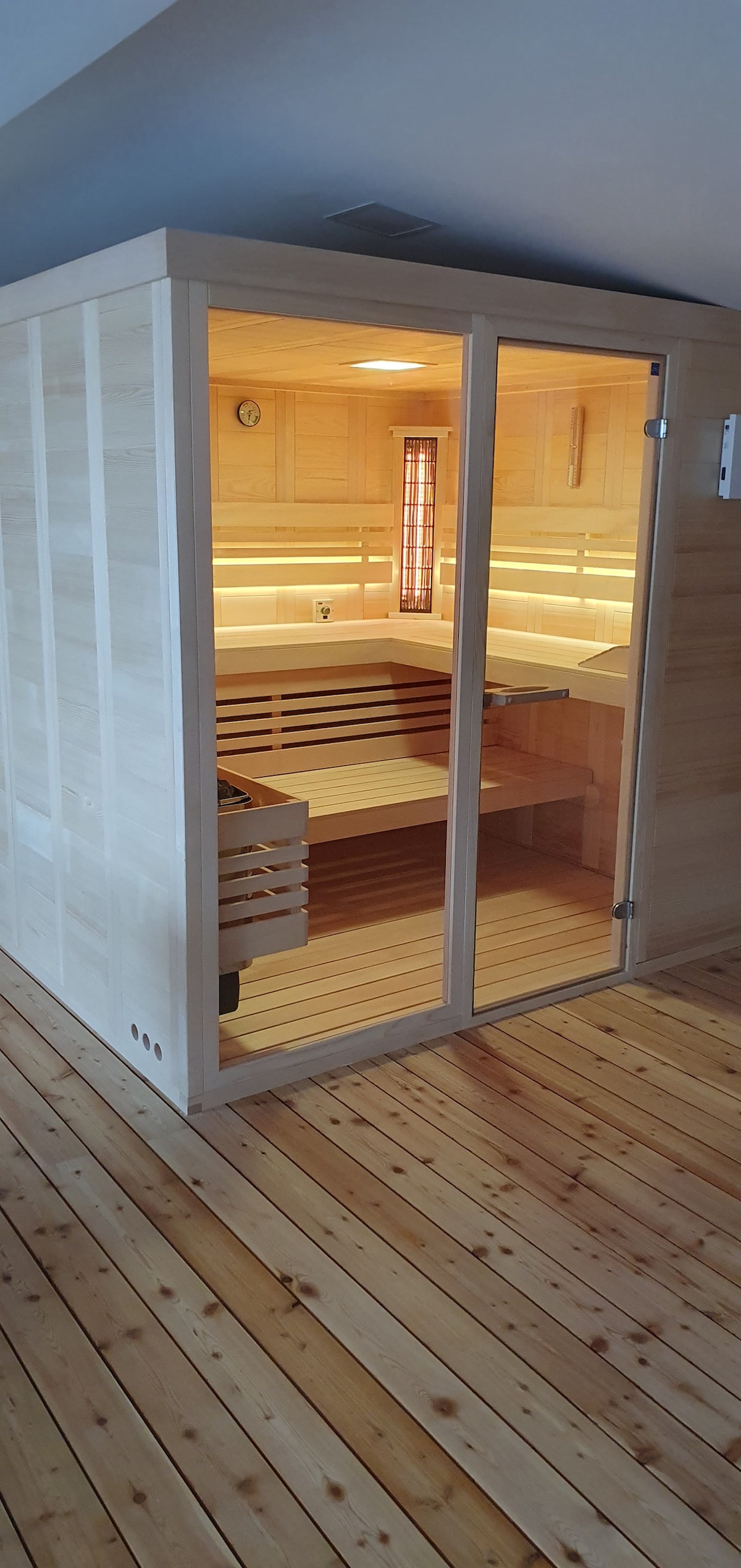 A wooden sauna is displayed with large glass doors, inviting light to fill the space. Interior benches made of light-colored wood are present, and warm lighting highlights the wooden panels, creating a serene atmosphere. A wooden floor complements the sauna's design.