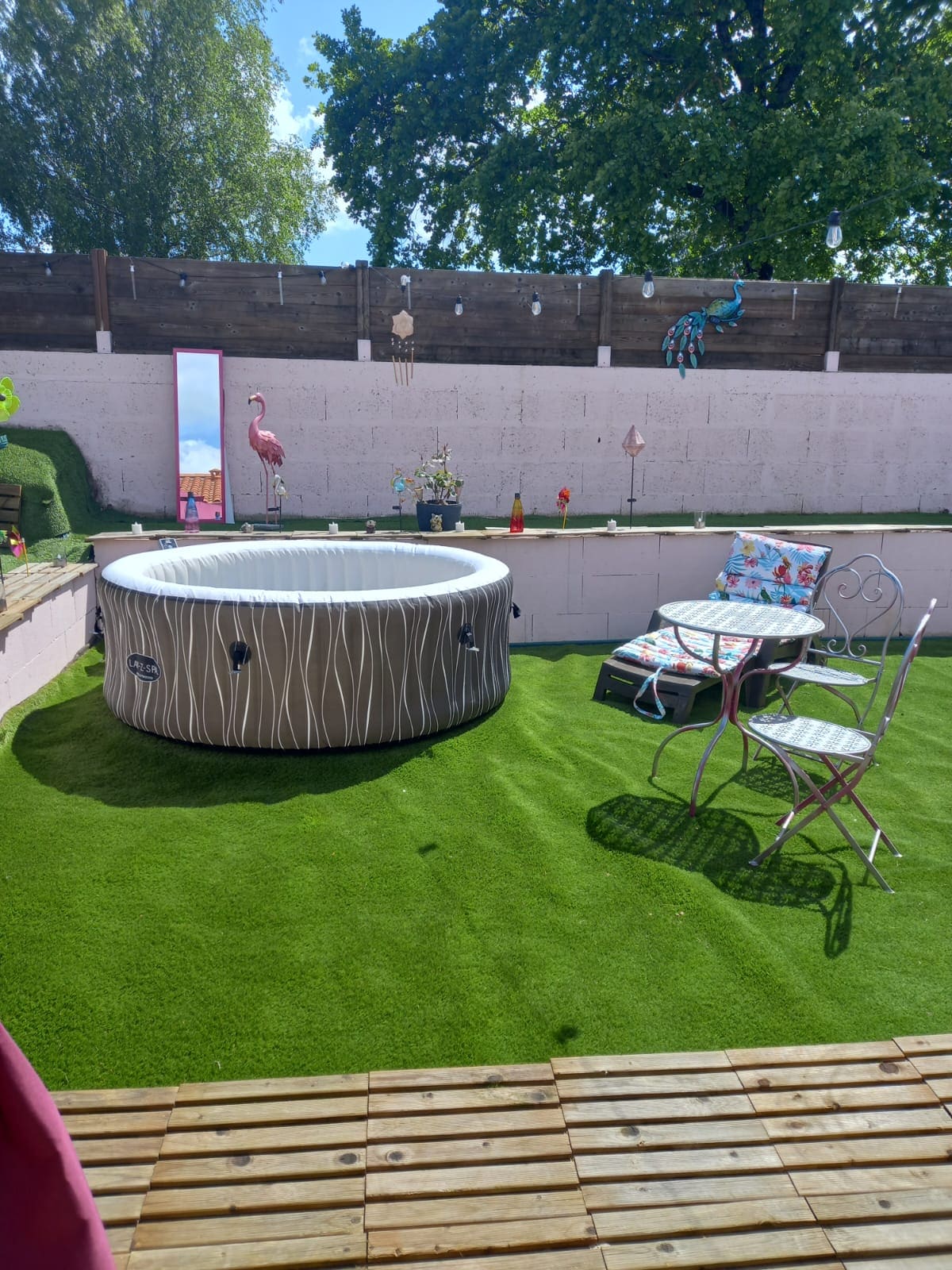 A spacious outdoor area is showcased, featuring a round inflatable spa surrounded by manicured green grass. A small table with stylish chairs invites relaxation, while colorful decor accents the space, including sculptures and a mirror positioned against the wall.