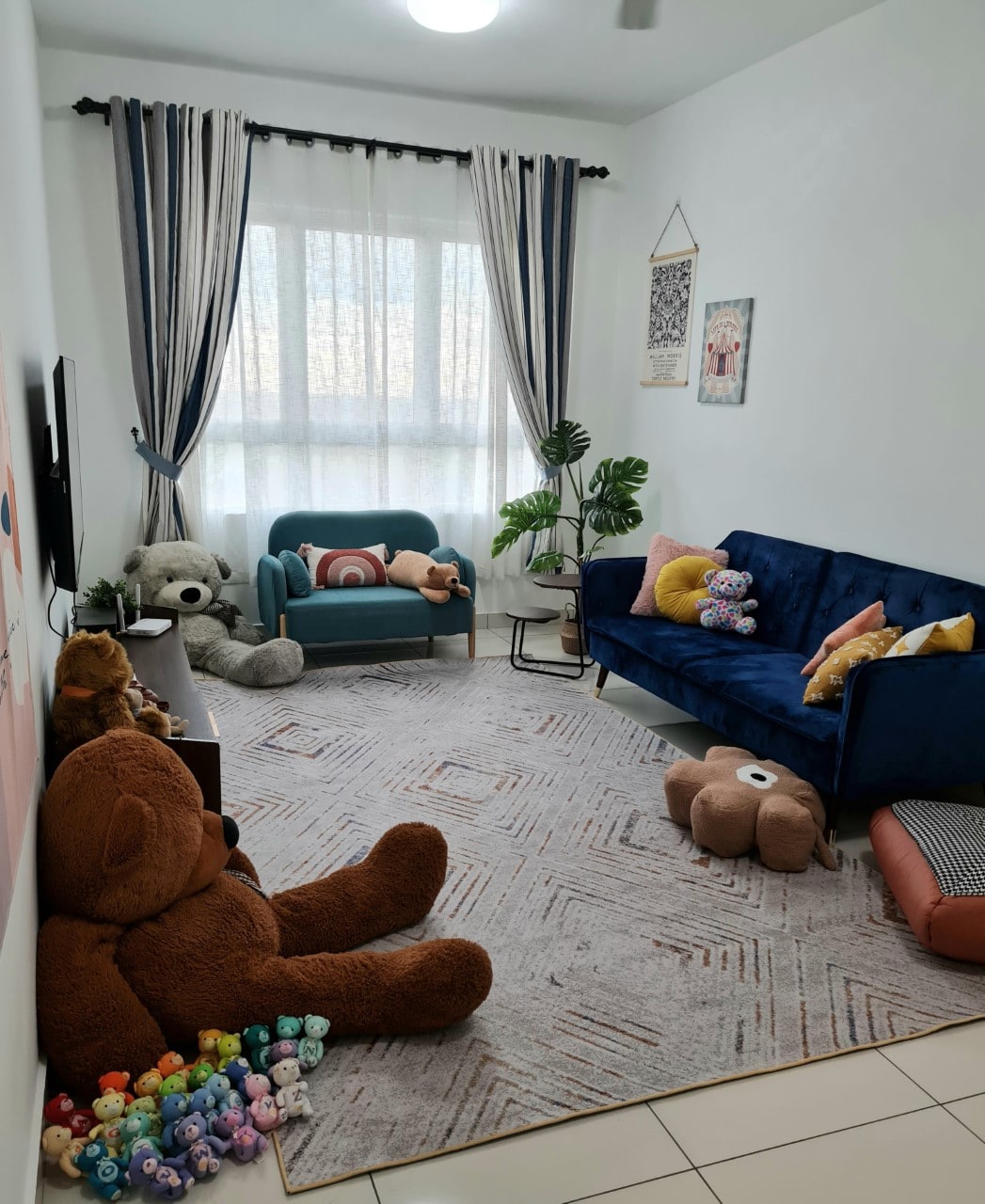 A cozy living area features a blue sofa and a chair, surrounded by soft toys and decorative pillows. A large window allows natural light to fill the space, accented by curtains with vertical stripes. A plush rug covers the floor, adding warmth to the room.