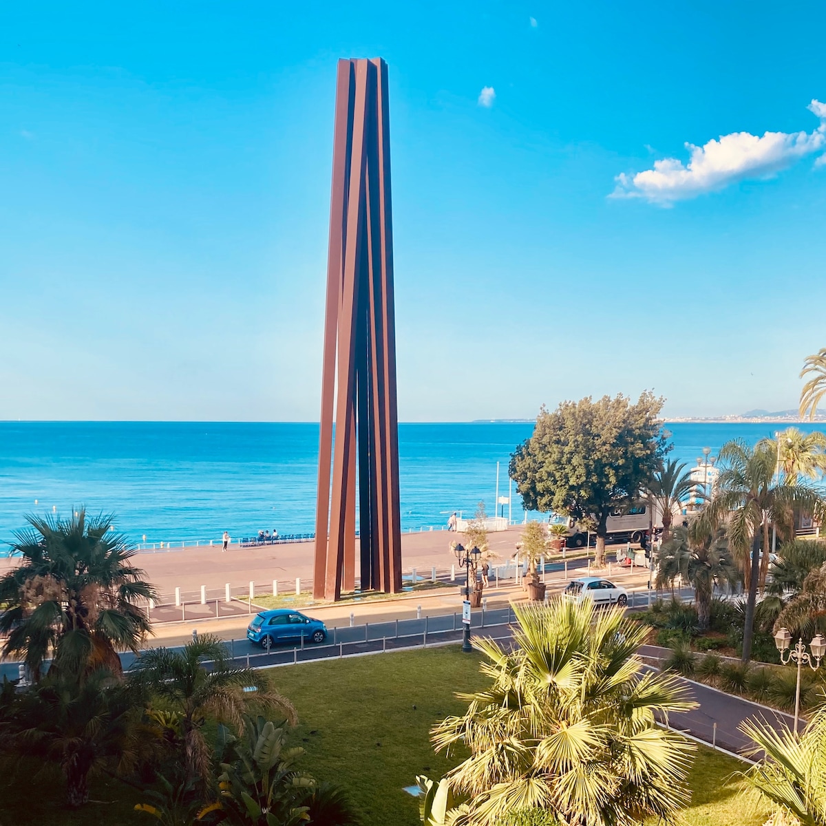 The image portrays a sculpture of metal beams situated facing the sea, with a clear blue sky above. Lush palm trees and greenery are visible in the foreground, alongside a road where cars and pedestrians can be seen. The tranquil blue waters of the bay stretch along the horizon.