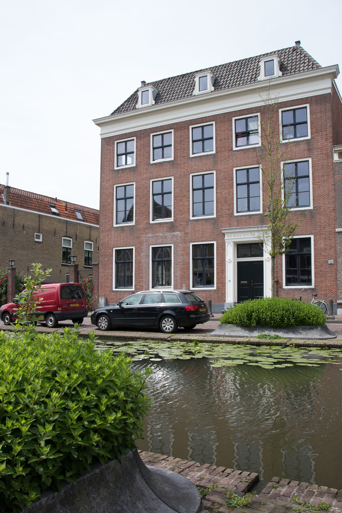 A historic canal house is presented with a red brick facade and multiple large windows. A tree grows near the entrance, while a peaceful canal lined with lily pads runs in front. Vehicles are parked along the street, contributing to the urban environment.